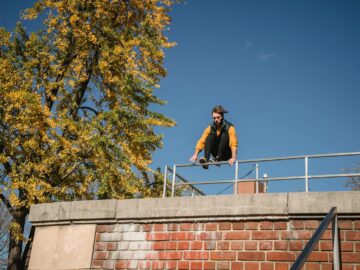 A young man executing a dynamic parkour jump over a brick wall in an urban setting, under a clear blue sky.