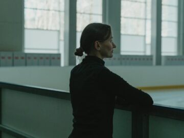 A woman in black clothing stands thoughtfully by an ice rink indoors.