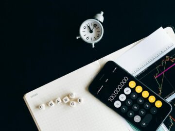 Notebook with an iPhone displaying 100,000,000, desk clock and financial charts suggesting market analysis.