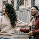A couple in casual clothing appears to be in a disagreement outdoors with grey fencing background.