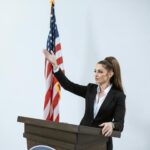 Confident woman in a black suit delivers a speech at a podium with an American flag backdrop.