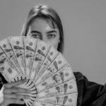 Young woman holding a fan of US dollar bills, partially covering her face, in a black and white portrait.