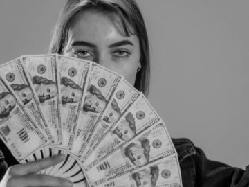Young woman holding a fan of US dollar bills, partially covering her face, in a black and white portrait.