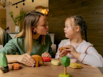 An adult woman and a child with Down syndrome engaging in playful interaction with wooden toys indoors.