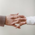 Crop unrecognizable female friends in casual outfit holding hands in light apartment near white wall
