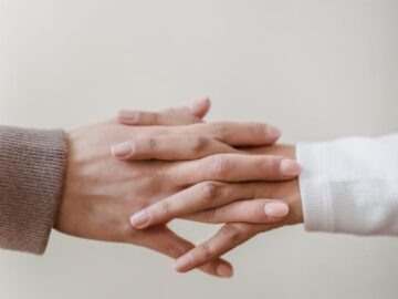 Crop unrecognizable female friends in casual outfit holding hands in light apartment near white wall