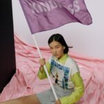 A young woman sits indoors holding a flag with 'Kindness' on it, embodying peaceful protest.