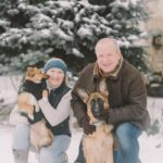 Senior couple with German Shepherd and another dog outdoors in snowy winter setting.