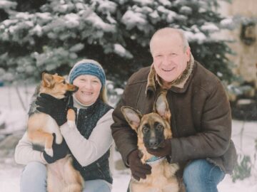 Senior couple with German Shepherd and another dog outdoors in snowy winter setting.