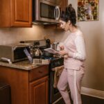 A woman in a kitchen cooking while referencing a laptop and notebook.