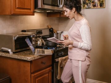 A woman in a kitchen cooking while referencing a laptop and notebook.