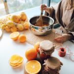 A woman stirring oranges in pot for homemade jam, with jars and fruits on the table.