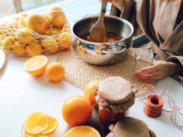 A woman stirring oranges in pot for homemade jam, with jars and fruits on the table.