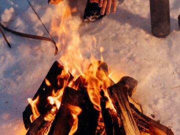 A person warms their hands near a campfire in a snowy winter scene, depicting warmth and coziness.