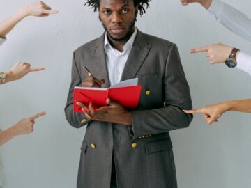 A man in a suit holding a red notebook with hands pointing at him, symbolizing pressure.