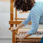 A woman in casual attire working with yarn on a wooden loom inside a workshop.