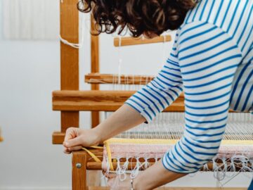 A woman in casual attire working with yarn on a wooden loom inside a workshop.