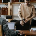 Young artisan selecting leather briefcase in workshop with laptop and numerous bags displayed in the background.