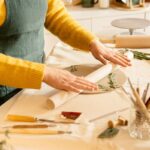A person in yellow sweater rolling clay with rosemary leaves on a work table indoors.