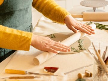 A person in yellow sweater rolling clay with rosemary leaves on a work table indoors.
