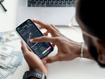 A person trading stocks using a smartphone, showcasing financial data on mobile and laptop screens.