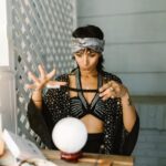 Mystical woman in patterned attire performing a ritual with crystal ball indoors.