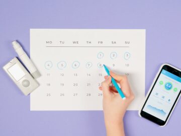 Hand marking days on a calendar with glucose monitor and smartphone app, emphasizing diabetes management.