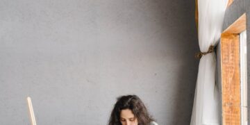 A young woman in casual attire painting on the floor of a modern studio with natural light.