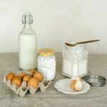 A rustic display of baking ingredients including eggs, milk bottle, and flour on a linen surface.