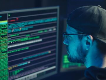 A cybersecurity expert inspecting lines of code on multiple monitors in a dimly lit office.