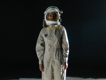 Astronaut standing in spacesuit on moon-like surface with black background.