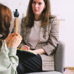 Serious adult female therapist in elegant outfit sitting in armchair and listening to crop anonymous patient talking about problems during psychotherapy consultation in light office