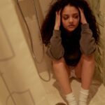 A young woman with curly hair sits thoughtfully in a shower, reflecting on her emotions.