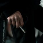 A close-up shot of a person holding a lit cigarette, depicting smoking habit indoors.