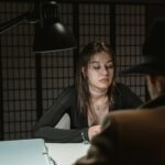 A woman and man sit across in a dimly lit interrogation room, suggesting mystery.