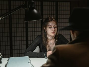 A woman and man sit across in a dimly lit interrogation room, suggesting mystery.