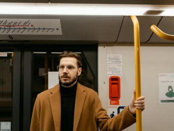 A man wearing a brown coat and glasses stands inside a subway train, holding a handrail.