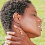 A woman outdoors holding her neck in discomfort, indicating pain or injury.