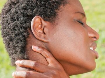 A woman outdoors holding her neck in discomfort, indicating pain or injury.