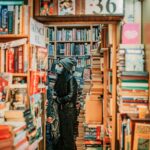 A young woman wearing a mask explores a bookstore filled with stacks of books.