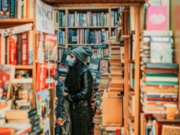 A young woman wearing a mask explores a bookstore filled with stacks of books.
