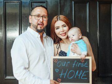 A joyful family stands at their new home's entrance holding a 'our first home' sign.