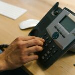 A hand dialing a phone on a wooden desk. Office environment with keyboard and planner.