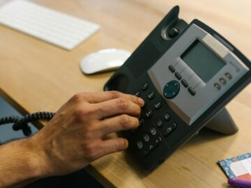 A hand dialing a phone on a wooden desk. Office environment with keyboard and planner.