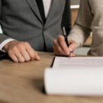 Close-up of businessmen signing documents at a wooden table in an office.
