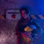 Young astronaut holding helmet in a dark emergency-themed room with red and blue lighting.