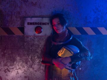 Young astronaut holding helmet in a dark emergency-themed room with red and blue lighting.