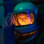 Portrait of a spacewoman in a helmet gazing intensely with colorful lighting.