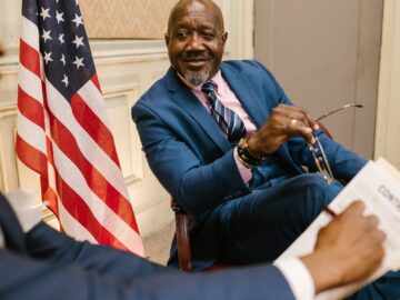 Two professionals in discussion by an American flag in an office setting.