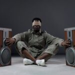 Blindfolded man seated with arms outstretched touching speakers on each side, exploring sound.